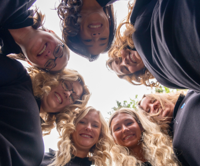 Group of seven people form a circle, heads together, looking down. They smile warmly, creating a sense of unity and joy. Outdoors with a cloudy sky.