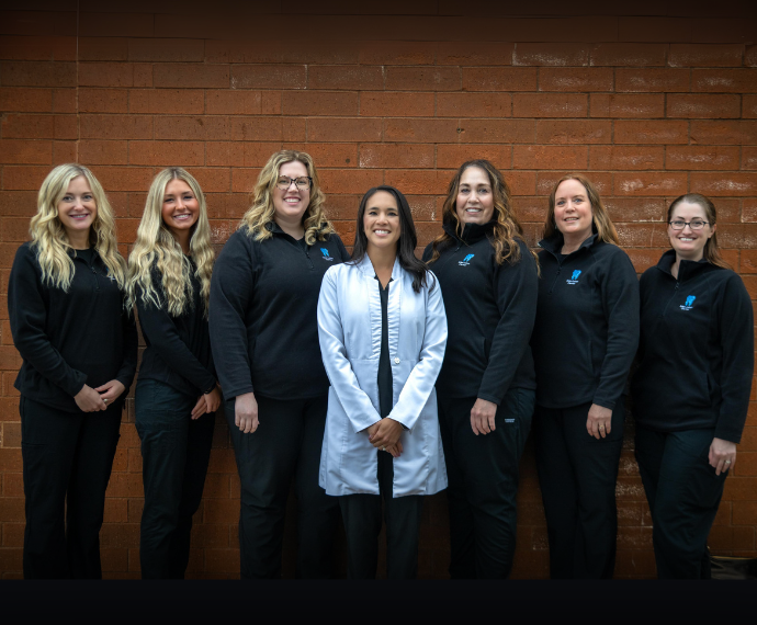 A group of seven women stands in front of a red brick wall, six wearing black uniforms, with one in the center in a white coat, all smiling warmly.