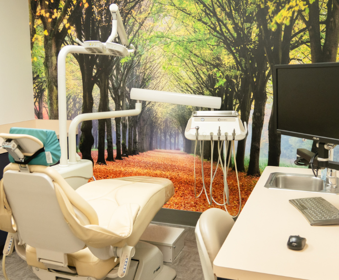 Dental office with a beige chair and equipment. A wall mural of a serene forest path with autumn leaves adds a calming, nature-inspired ambiance.