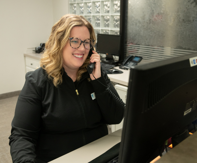 Smiling woman with glasses, wearing a black jacket, sits at a desk answering the phone beside a computer monitor in a modern office setting.