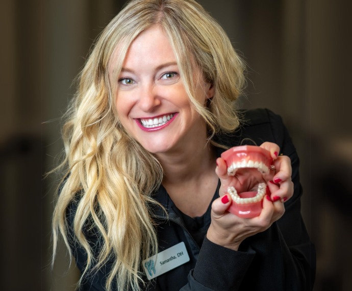 Smiling woman with long blonde hair holds a dental model, displaying teeth. She wears a black top and radiates a cheerful, professional demeanor.