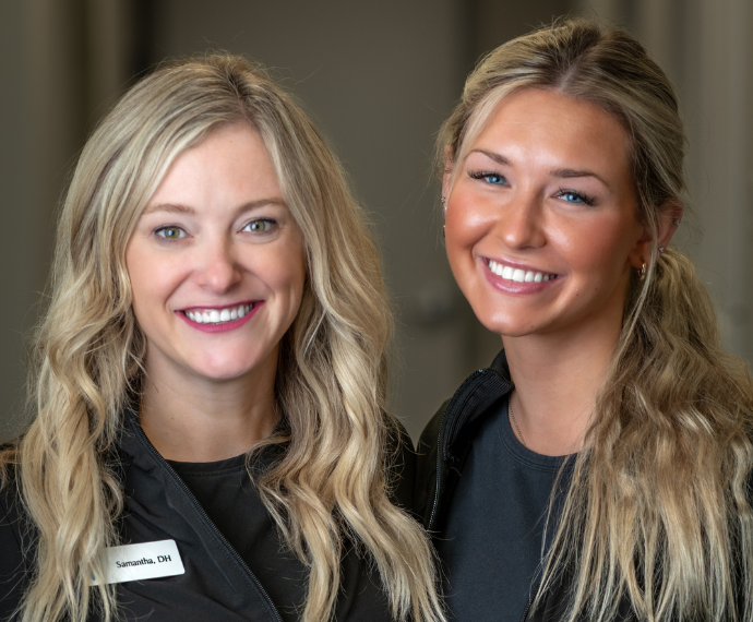 Two smiling women with long blonde hair stand closely together indoors, dressed in black attire. The tone is warm and friendly.