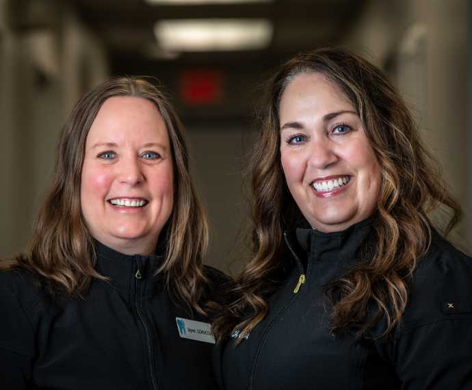 Two smiling women with long hair in black jackets stand in a hallway. They appear friendly and approachable, with bright overhead lighting.