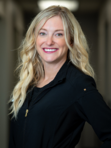 Smiling woman with long blonde hair wearing a black jacket, standing in a softly lit hallway. She exudes warmth and confidence.