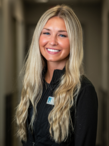 Smiling woman with long blonde hair in a black vest stands in a well-lit hallway. The atmosphere is friendly and welcoming, conveying warmth and openness.