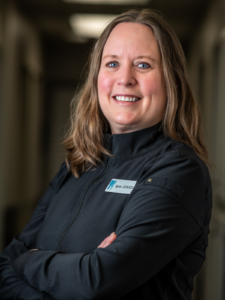 Smiling woman with long brown hair stands confidently with arms crossed, wearing a black jacket with a name tag in an office hallway.