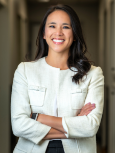 Smiling woman with long dark hair stands confidently with arms crossed, wearing a white blazer in a hallway. The tone is professional and warm.