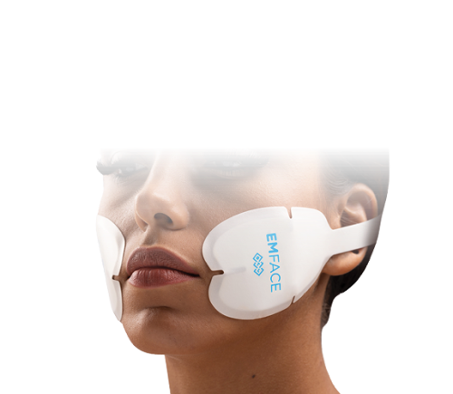 A person wears a futuristic face mask device labeled "EMFACE" on their cheeks, set against a black background. The mood is clinical and innovative.