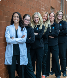 A diverse group of six women stand side by side against a brick wall, smiling. The first woman wears a white coat, while the others wear matching black outfits.