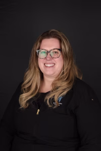 Smiling woman with curly blonde hair in a black zip jacket, wearing a name badge. She stands against a neutral background, exuding confidence.